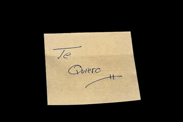 Yellow adhesive paper, similar to a Post-it note, on a black background with the word “Te Quiero”  in Spanish handwritten in blue ink.