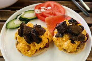 close up of American breakfast with scramble egg on muffin bread in half and stir fried mushroom. uncooked tomato and cucumber on the side on a white plate on a wooden table