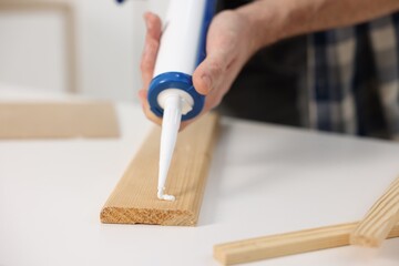 Man with caulking gun glueing wooden plank at white table, closeup