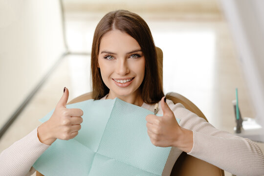Smiling beautiful girl is sitting in dentistry chair with positive emotion and gesturing fingers up. Female patient visit dentist for regular teeth mouth cavity check up at medical clinic
