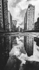 Fototapeta premium Black and white cityscape with reflections in puddle, showcasing urban architecture and dramatic clouds