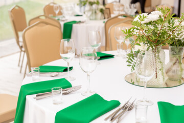 Close up of wine glasses, silver tableware and green napkins on round tables covered with white tablecloth.