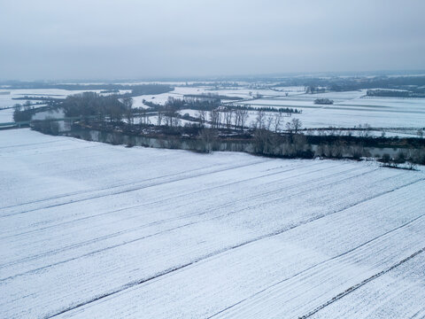 Aerial view of a snowy landscape showcasing vast fields and bare trees under a gray sky. Calm and serene winter scene.