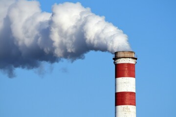 White smoke is coming out of a large red and white industrial chimney against a clear blue sky, symbolizing pollution and environmental concerns