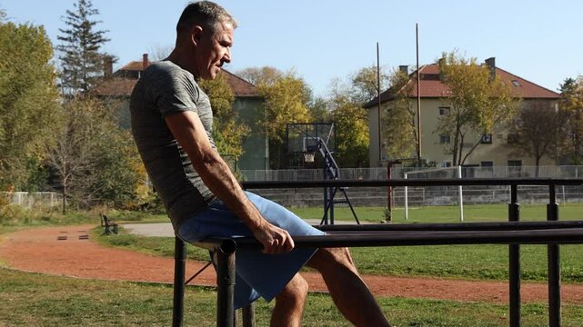 54-Year-Old Fit Man Doing Crunches - Athletic Track Background. Man in excellent physical condition doing crunches on parallel bars. Suitable for promoting fitness and strength training.