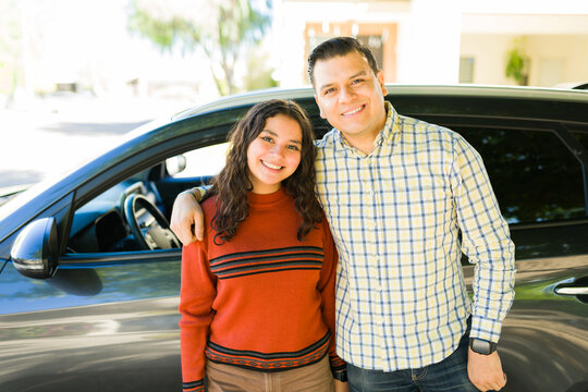 Proud father teaching his teenage daughter driving lessons, standing next to their car and smiling