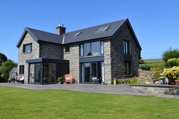 This contemporary stone house boasts a spacious lawn and patio area, beautifully situated in a peaceful countryside setting on a bright, sunny day