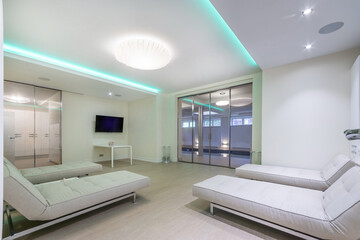 A relaxation area in a luxurious spa center. Lounge chairs with soft mattresses, a large .TV on the wall, a glass entrance door to the pool.