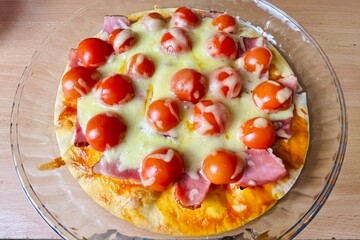 On a large glass plate lies a homemade gluten-free pizza with bacon, cherry tomatoes and cheese. homemade pizza
