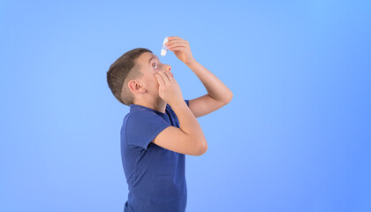 Side view of boy suffering from dry eyes applying eye lubricant while standing over blue background