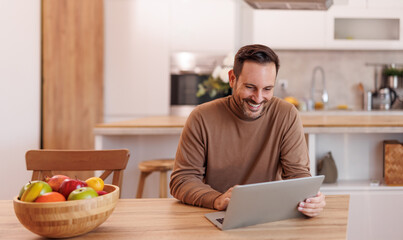 Happy mid adult male entrepreneur reading e-mails over laptop on dining table while working at home
