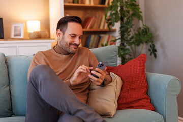 Handsome entrepreneur smiling and messaging over mobile phone while relaxing on sofa in living room