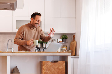 Happy mid adult man with coffee mug waving hand over video call on laptop while working from home