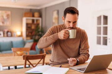 Portrait of mid adult businessman drinking coffee while working online over laptop on desk at home