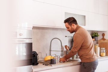 Side view of handsome mid adult man washing utensils under running water in kitchen sink at home