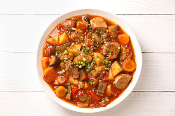 Delicious stew with vegetables in bowl on white wooden table, top view