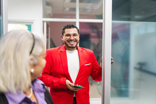 A cheerful man in a bright red blazer stands near a glass door, holding a smartphone