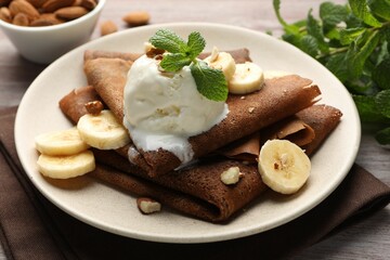 Delicious chocolate crepes with banana, nuts, mint and scoop of ice cream on wooden table, closeup