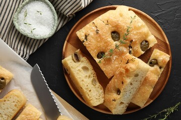Slices of delicious focaccia bread with olives, thyme, salt and knife on black table, flat lay
