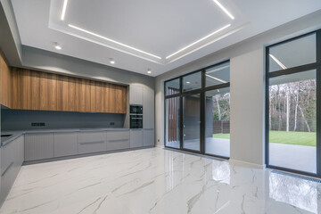 Modern kitchen design in a country house with panoramic windows. Grey kitchen set with built-in appliances. White tiled floor.