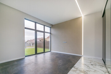 The room is unfurnished, with a panoramic window. Tile floor. Courtyard with a green .lawn outside the window of a country house.