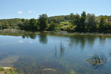 A peaceful scene of the Dobra River near Jaskovo, showcasing its clear waters reflecting the sky and surrounding greenery, a small dam, and an old bridge in the distance