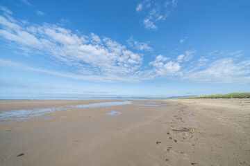 Expansive view of scenic Harlech Beach dunes and grass and patterns in sand to waters edge and horizon under blue sky with high wind-blown white cloud.