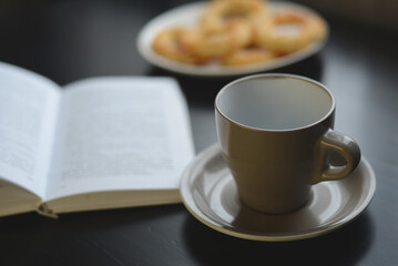 A cup of coffee, now empty, sits beside an open book on a dark surface, with a plate of cookies in the background. A cozy scene of taking a break from reading.