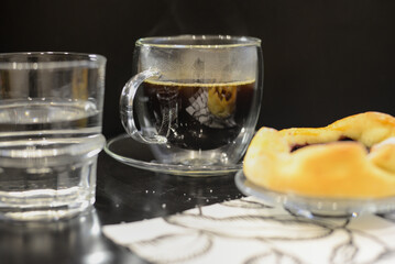 A clear glass mug of steaming coffee, a glass of water, and a golden-brown pastry with a dark filling on a plate, set on a dark table with a white napkin