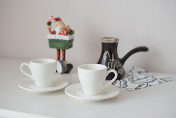 Two white coffee cups and saucers on a white table with a black coffee pot labeled 