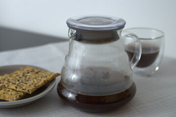 A glass carafe filled with coffee, a clear glass of coffee with a handle, and a plate of seed-covered crackers, all set on a white tablecloth for a cozy and inviting scene