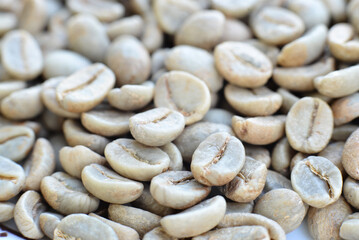 A close-up shot of pale green, unroasted coffee beans with a slightly wrinkled texture. The beans are arranged randomly, with some in focus and others blurred, showcasing their natural form.
