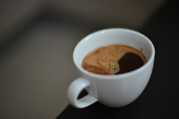 A close-up shot of a white coffee cup filled with dark coffee, topped with a thin layer of foam, resting on a dark surface for a simple and inviting look