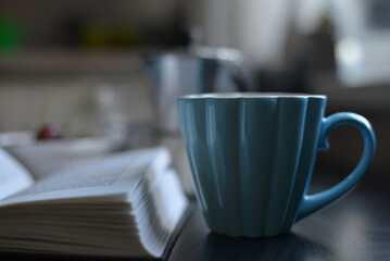 A blue mug with a fluted design and heart-shaped handle sits on a dark surface next to an open book. The blurred background suggests a cozy, intimate setting perfect for relaxation.