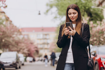 Young businesswoman using smartphone while walking down the street in spring