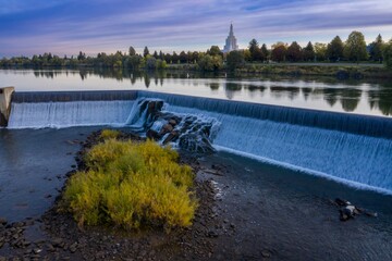 Tranquil river scene with a dam cascading water, reflecting the serene sky and distant temple. Peaceful autumn colors. Snake River, Idaho Falls, Idaho, USA.