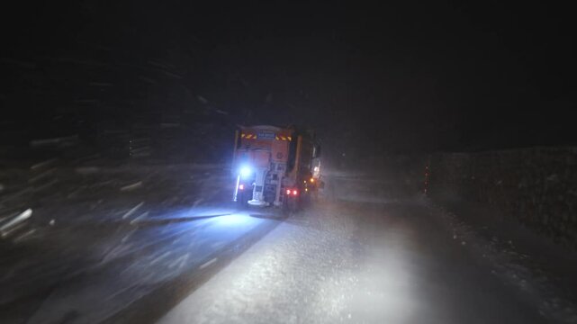A snowplow is working on a snow-covered road at night, illuminating the path with its lights. Barriers and directional signs are visible on the side. The vehicle ensures the road remains clear safe.