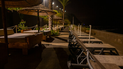 A quiet beachfront lounge area at night with empty sunbeds, straw umbrellas, and soft ambient lighting. Palm trees and a calm atmosphere create a tranquil seaside retreat.