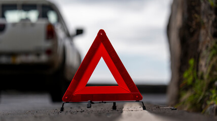 A red warning triangle placed on a roadside, with a parked white car in the background. The scene highlights a roadside safety precaution in a natural outdoor setting.