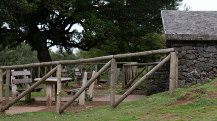 A rustic picnic area with wooden tables, benches, and a wooden fence, set near a stone building and surrounded by lush trees. The scene exudes a peaceful, countryside atmosphere.