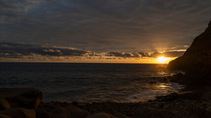 Obraz premium A dramatic sunset over the ocean, with golden light reflecting on the water and dark rocky cliffs framing the scene. A serene and atmospheric coastal landscape.
