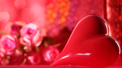 A shiny red heart stands out against a bokeh-filled background, with soft pink roses softly blurred, offering a heartfelt depiction of love and affection.