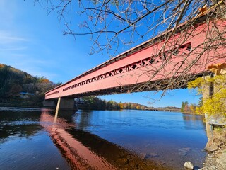 Wooden red covered bridge extending across a calm river with blue water and reflected in the water's surface.