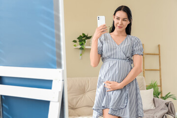 Young pregnant woman in beautiful dress with mobile phone taking picture near mirror at home