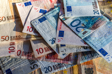 Euro banknotes of diverse denominations spread over a table, seen at an angle, exposed to the sun light with shadows. 5, 10, 20 and 50 bills visible.