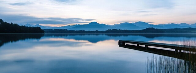 A serene view of a calm lake at dusk, with a small wooden pier extending into the water and distant...