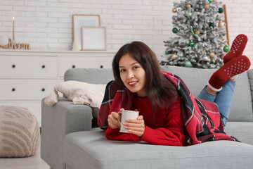 Beautiful young happy Asian woman with cup of hot mulled wine lying on sofa near Christmas tree at...