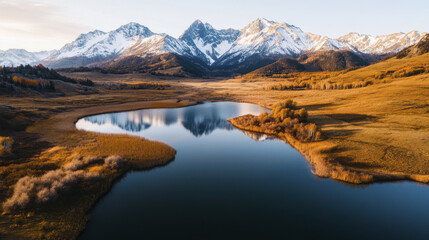 Obraz premium Aerial view of tranquil lake reflecting snow capped mountains, surrounded by golden grasslands and autumn foliage, creating serene landscape