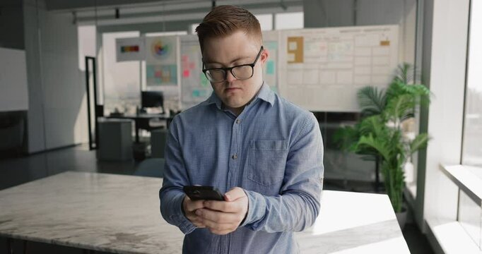 Man with Down syndrome hold smartphone standing in modern workplace, manage tasks, using apps to organize daily schedule, set reminder, respond to work-related messages, share updates with colleagues