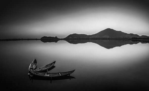 In the stillness of a black-and-white world, a few wooden boats are moored on the shore of a placid lake